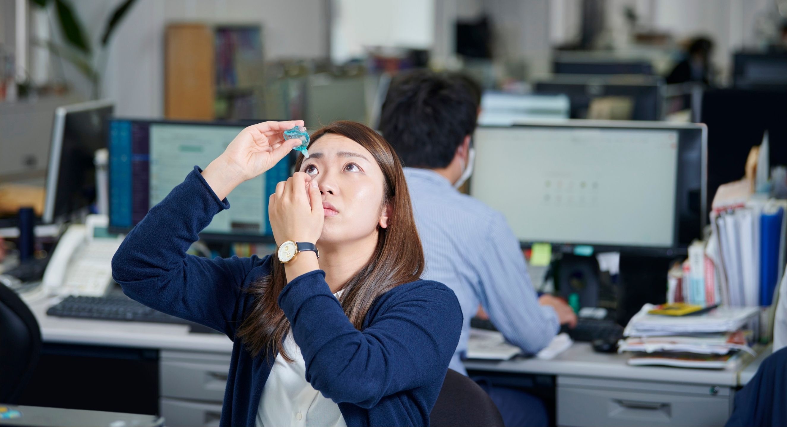 Person at desk in busy office self-administering eye drops