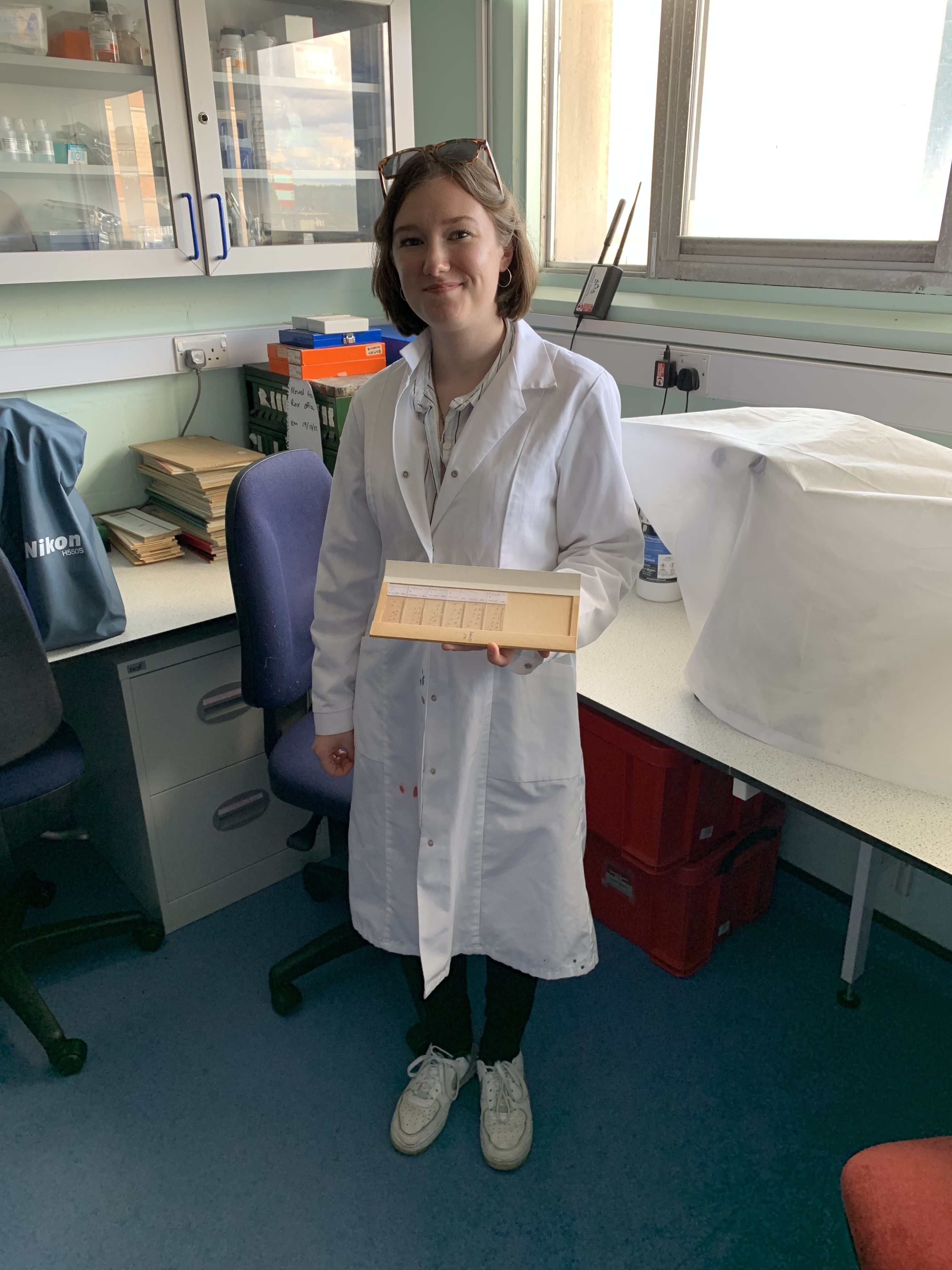 a PhD student standing in their laboratory wearing a white lab coat and smiling at the camera